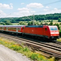 Ein roter Regionalzug der Deutschen Bahn mit einer Elektrolokomotive und mehreren Personenwagen fährt mit hoher Geschwindigkeit auf einer mehrgleisigen Bahnstrecke. Die Aufnahme zeigt den Zug von schräg vorne in einer dynamischen Vorbeifahrt, was durch eine leichte Bewegungsunschärfe an den Gleisen und dem unteren Bereich des Zuges betont wird. Im Hintergrund erstreckt sich eine hügelige, grüne Landschaft mit Wäldern und Feldern unter einem leicht bewölkten, blauen Himmel. Das Bild symbolisiert Mobilität und Schienenverkehr als Ergänzung zu anderen Reiseformen.